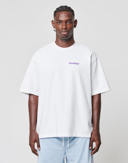 A man faces forward against a light gray background, wearing the LIVE FAST DIE YOUNG Basic 280 Tee—an oversize white t-shirt made of 100% Baumwolle with LiveFast in small purple text on the chest—and light blue shorts.