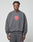 A person stands facing forward in a loose-fitting DY Sweater by LIVE FAST DIE YOUNG, made from cotton. The dark grey oversize sweatshirt displays a red and white graphic logo on the chest. The background is plain and light-colored.