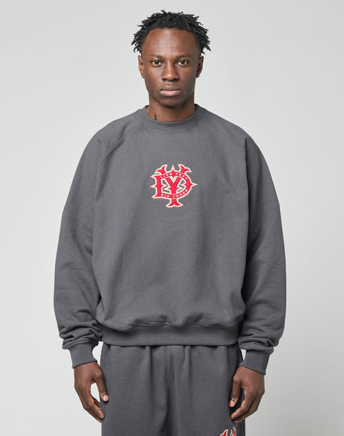 A person stands facing forward in a loose-fitting DY Sweater by LIVE FAST DIE YOUNG, made from cotton. The dark grey oversize sweatshirt displays a red and white graphic logo on the chest. The background is plain and light-colored.