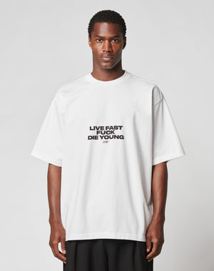 A person stands facing forward in the LIVE FAST DIE YOUNG Fuck Die Young Tee, a loose white T-shirt with an edgy front print, set against a plain light gray background.