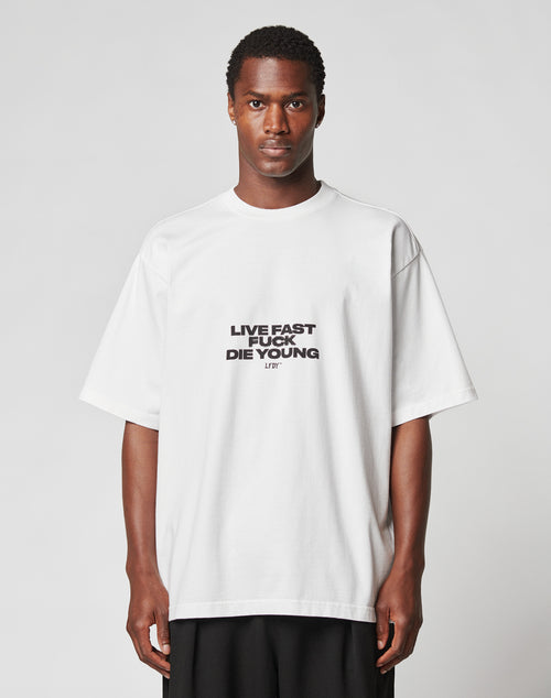 A person stands facing forward in the LIVE FAST DIE YOUNG Fuck Die Young Tee, a loose white T-shirt with an edgy front print, set against a plain light gray background.
