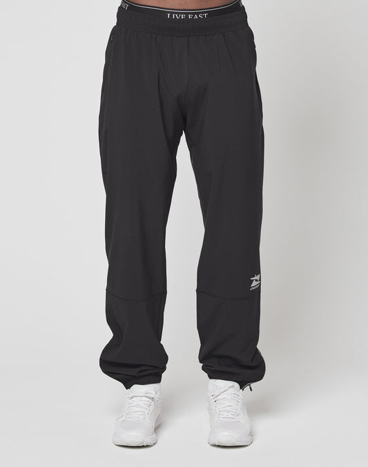A person wears LIVE FAST DIE YOUNGs Live Fast Performance Running Pants—black joggers with a reflective print by the left knee, an elastic waistband for comfort—and white sneakers, standing against a plain white background.