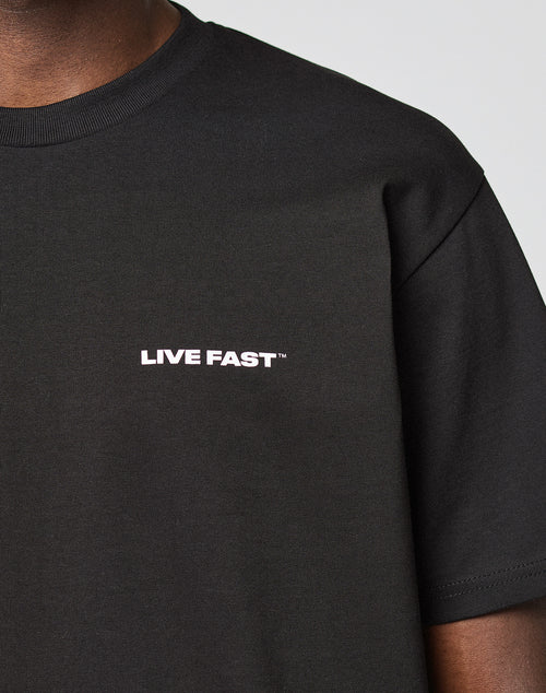 A close-up shows a person wearing the LIVE FAST DIE YOUNG Live Fast Plain Tee, a black Regular Fit t-shirt with “LIVE FAST™” in bold white letters on the chest. Made from 100% Baumwolle; their face is not visible against a plain background.