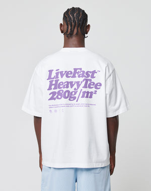 A person with braided hair is seen from behind, wearing a white Basic 280 Tee by LIVE FAST DIE YOUNG with purple Front- und Backprint text and smaller details, paired with light blue shorts.
