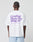 A person with braided hair is seen from behind, wearing a white Basic 280 Tee by LIVE FAST DIE YOUNG with purple Front- und Backprint text and smaller details, paired with light blue shorts.