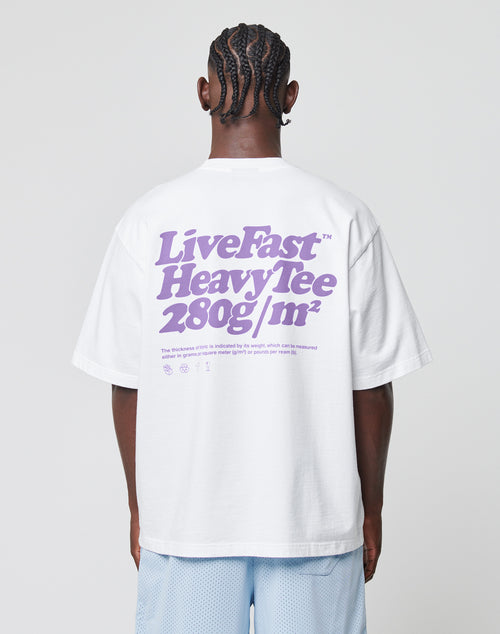A person with braided hair is seen from behind, wearing a white Basic 280 Tee by LIVE FAST DIE YOUNG with purple Front- und Backprint text and smaller details, paired with light blue shorts.