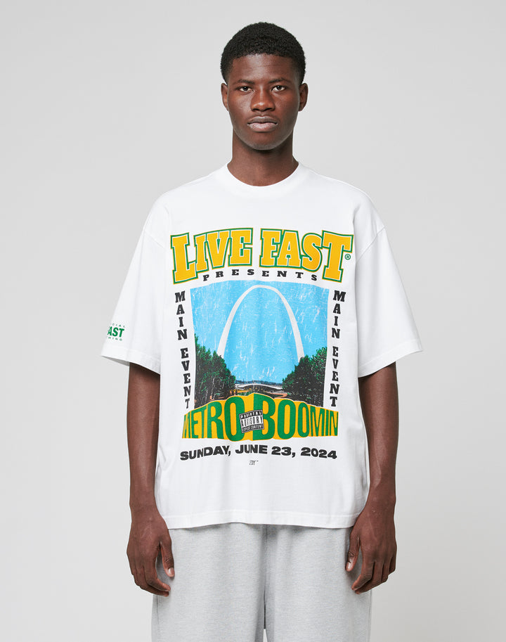 A man faces forward in a white LIVE FAST DIE YOUNG LFDY Metro Boomin Tee featuring a colorful front print with event details, paired with light gray pants against a plain light gray background. | model