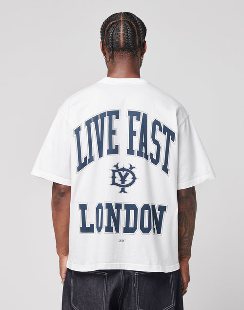 A person with braided hair faces away, wearing the LIVE FAST DIE YOUNG Live Fast London Tee, a white oversized 100% Baumwolle T-shirt featuring bold blue LIVE FAST LONDON and a circular logo on the back, paired with dark jeans.