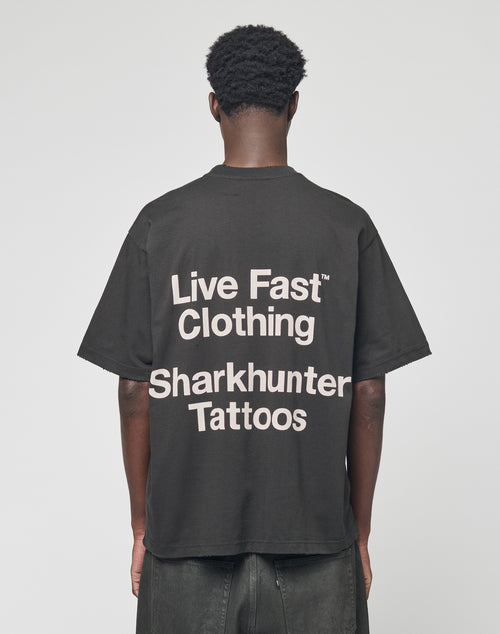 A person stands with their back to the camera, wearing the LFDY Sharkhunter Tee by LIVE FAST DIE YOUNG, an oversized black cotton T-shirt featuring bold white Sharkhunter Tattoos text on the back.