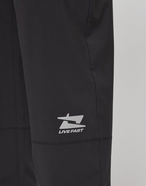 Close-up of Live Fast Performance Running Pants by LIVE FAST DIE YOUNG. Black, oversize fit with a white stylized bird/arrow logo and LIVE FAST text in reflektierender Print on the fabric.