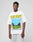 A man wearing the LFDY Metro Boomin Tee by LIVE FAST DIE YOUNG, featuring an oversize white cotton fit, bold yellow LIVE FAST print, blue and green graphic, and event details, stands against a plain gray background.