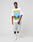 A man stands in a studio facing forward, wearing the LIVE FAST DIE YOUNG LFDY Metro Boomin Tee in an oversize fit with front print, paired with light gray knee-length shorts, white socks, and sneakers. Made from soft Baumwolle. Plain gray background.