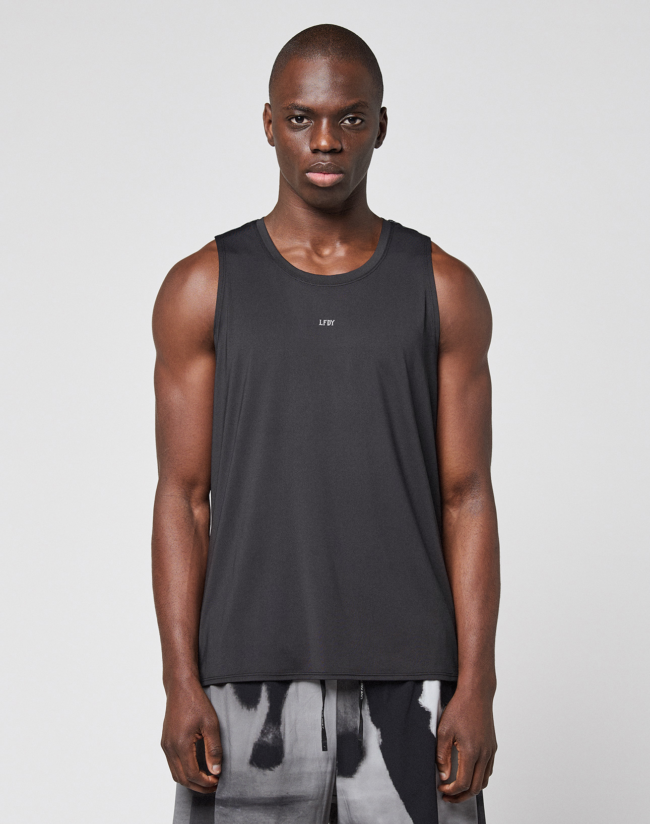 A man with a neutral expression stands against a plain background wearing the LIVE FAST DIE YOUNG LFDY Performance Tank, a regular fit black sleeveless athletic shirt with reflective logo print, paired with black-and-white patterned shorts.