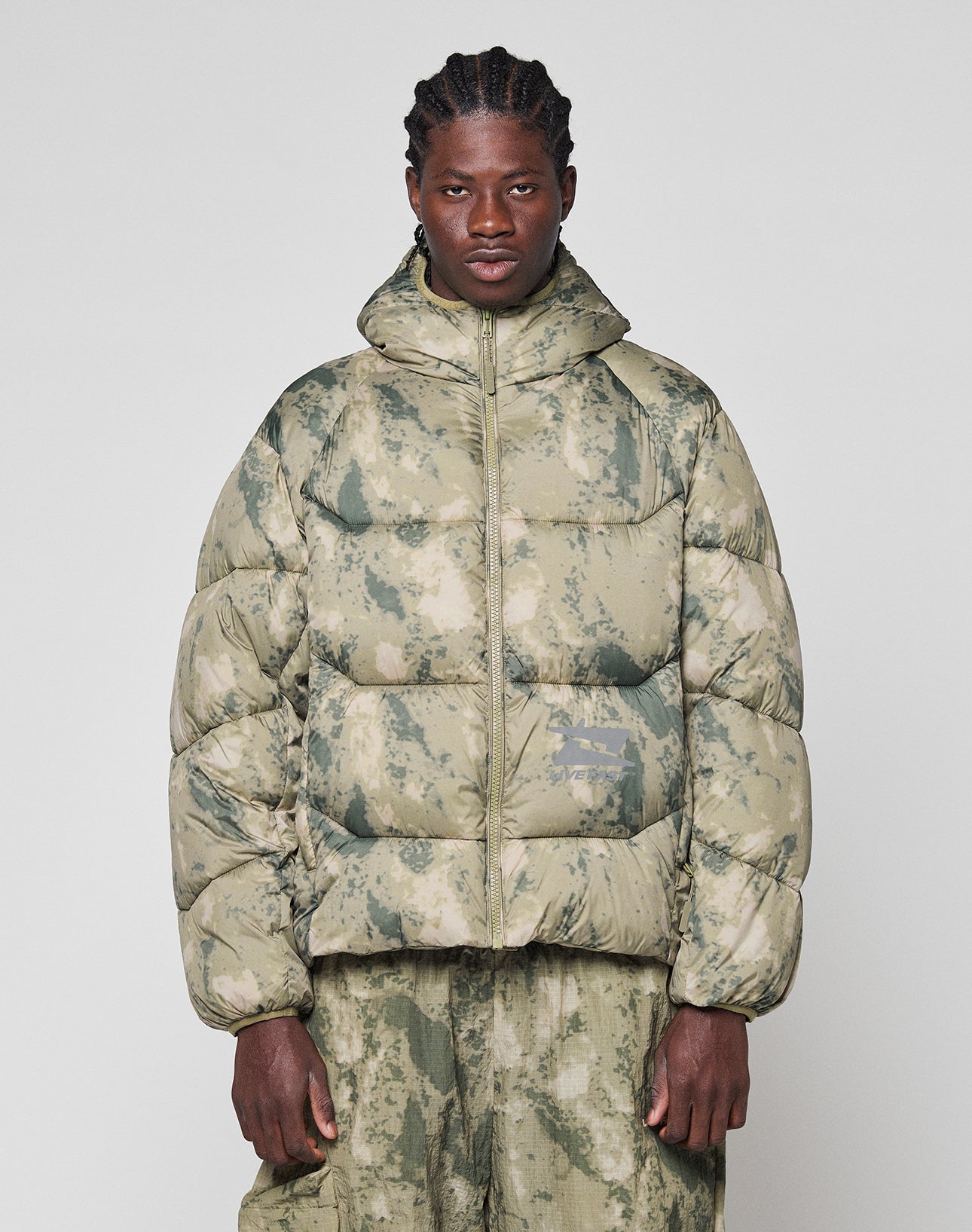 A person with braided hair wears the LIVE FAST DIE YOUNG Live Fast Puffer Jacket in green and beige camouflage with matching oversize-fit pants, standing against a plain light background.