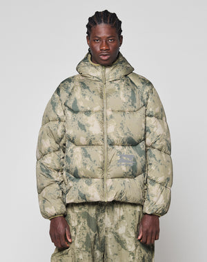 A person with braided hair wears the LIVE FAST DIE YOUNG Live Fast Puffer Jacket in green and beige camouflage with matching oversize-fit pants, standing against a plain light background.
