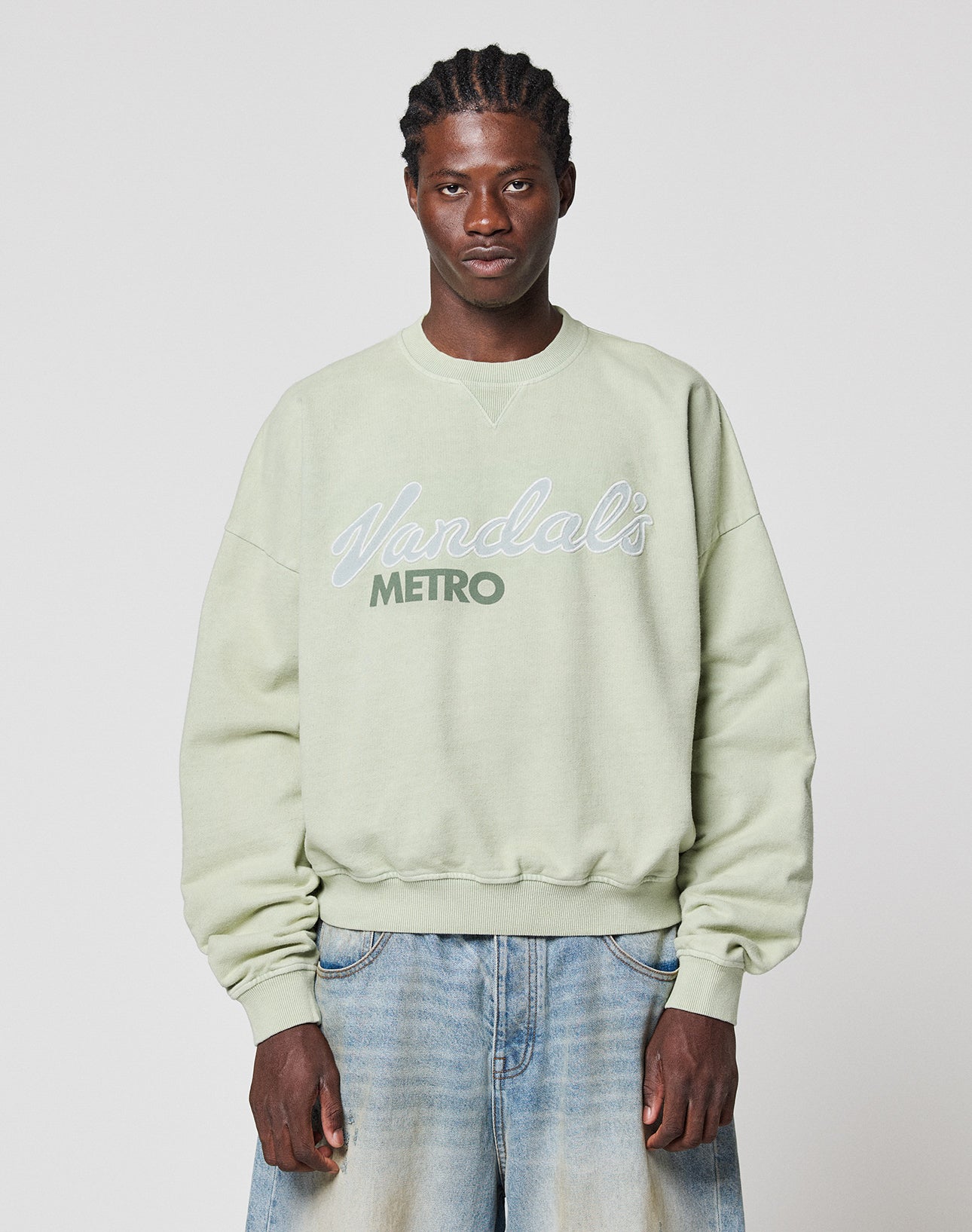 A man with braided hair wears a light green LFDY Vandals Sweater by LIVE FAST DIE YOUNG in Oversize Fit and light blue baggy jeans, standing against a plain white background.