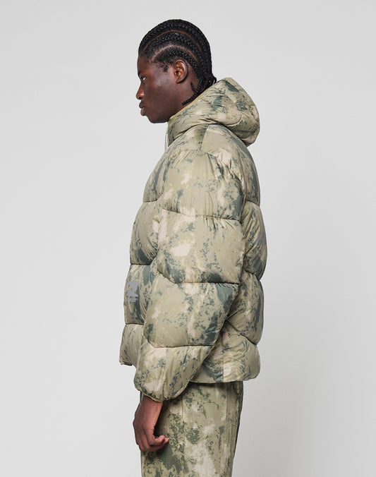 A person with braided hair stands sideways, wearing the LIVE FAST DIE YOUNG Live Fast Puffer Jacket—an oversize, green and beige camo hooded jacket with matching pants—against a plain light background.