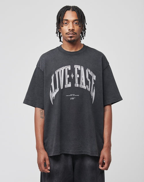 A man with braided hair faces forward, wearing a LIVE FAST DIE YOUNG Live Fast Axe Tee—an oversize black cotton T-shirt featuring a bold white LIVE FAST front print. The background is plain and light-colored.