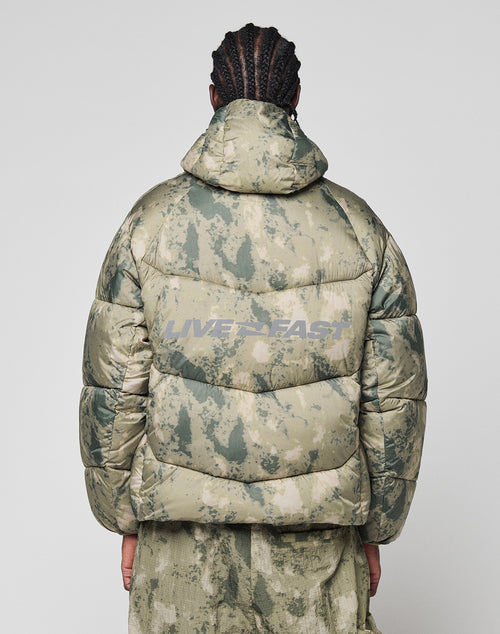 A person with braided hair, seen from behind, wears the LIVE FAST DIE YOUNG Live Fast Puffer Jacket—an oversized green camo jacket with hood and bold gray reflective "LIVE FAST" print on the back.