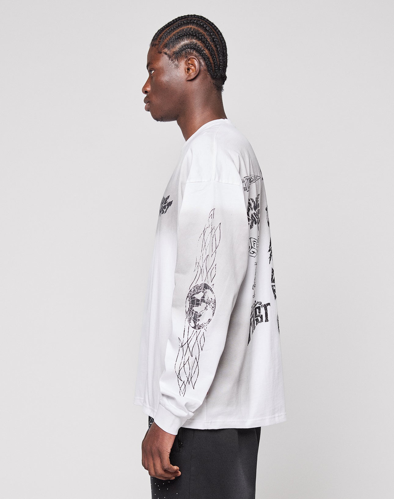 A person with braided hair stands in profile wearing the LIVE FAST DIE YOUNG LFDY Logo Longsleeve, a white 100% cotton shirt with black graphics on the back and sleeves, plus black pants, against a plain light gray background.