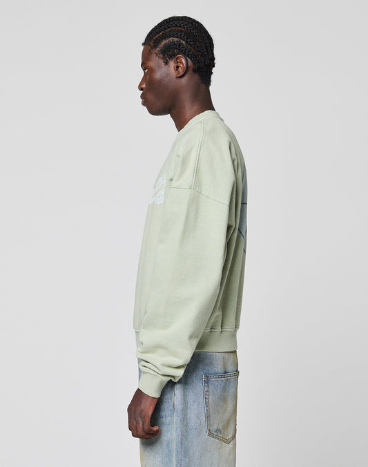 A person with braided hair stands in profile against a plain light gray background, wearing the LIVE FAST DIE YOUNG LFDY Vandals Sweater in light green and light blue jeans.