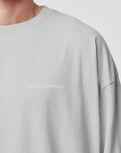 A close-up of a person wearing an oversize fit, light gray LIVE FAST DIE YOUNG Basic Longsleeve made from 100% Baumwolle, featuring a subtle logo print on the left chest. Only part of the neck and shoulder is visible.