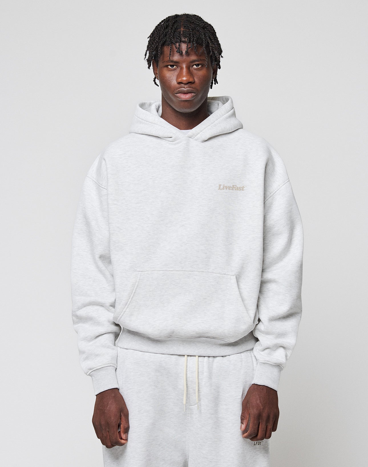 A person with short, twisted hair wears the LIVE FAST DIE YOUNG Basic 360 Hooded in light gray and matching sweatpants, standing against a plain light background and looking directly at the camera.
