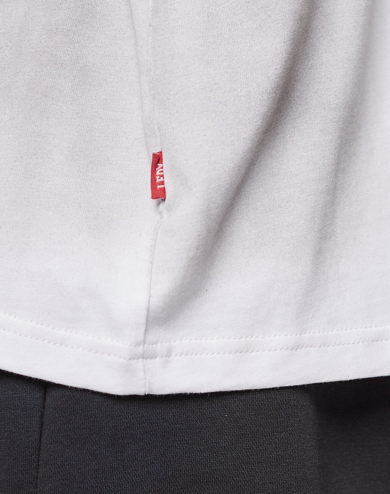 Close-up of the lower side of a white LFDY Logo Longsleeve by LIVE FAST DIE YOUNG, Oversize Fit, featuring a small red fabric tag sewn into the seam above black pants.