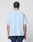 A person with neatly braided hair is shown from the back, wearing the LIVE FAST DIE YOUNG Live Fast College Tee in oversize light blue 100% Baumwolle and navy shorts with white accents, standing against a plain light gray background.