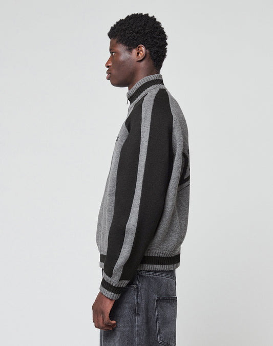 A person stands in profile against a plain background, wearing the LIVE FAST DIE YOUNG Boxy Knit Zip Jacket with gray and black vertical-striped sleeves, paired with dark gray jeans.