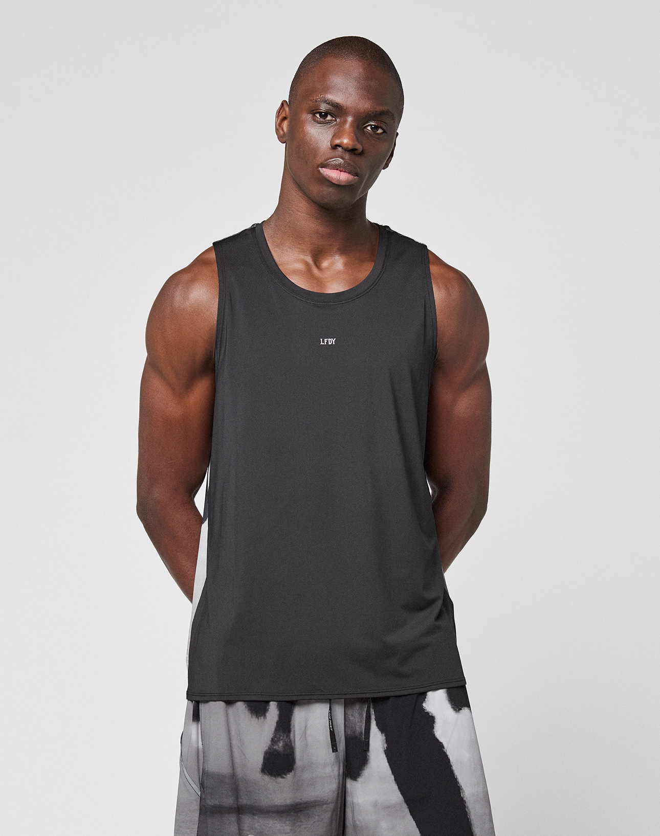 A man stands against a plain light background wearing the LIVE FAST DIE YOUNG LFDY Performance Tank, featuring leichtes Obermaterial, and black-and-white patterned shorts. His arms are relaxed behind his back with a neutral expression.