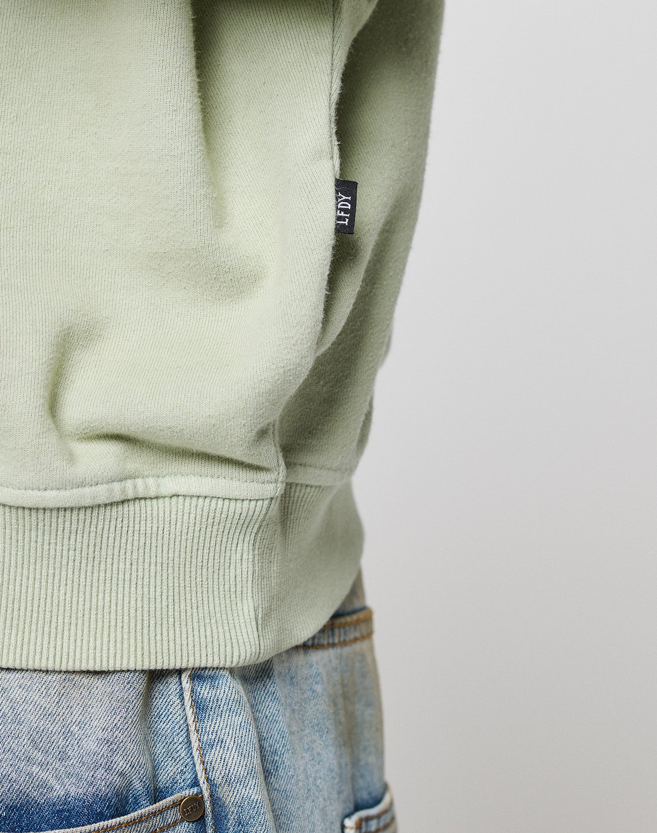 Close-up of a person wearing the LIVE FAST DIE YOUNG LFDY Vandals Sweater in light green, featuring a small black tag and partially tucked into light blue jeans. Crafted from 100% Baumwolle for comfort. Background is plain and light-colored.