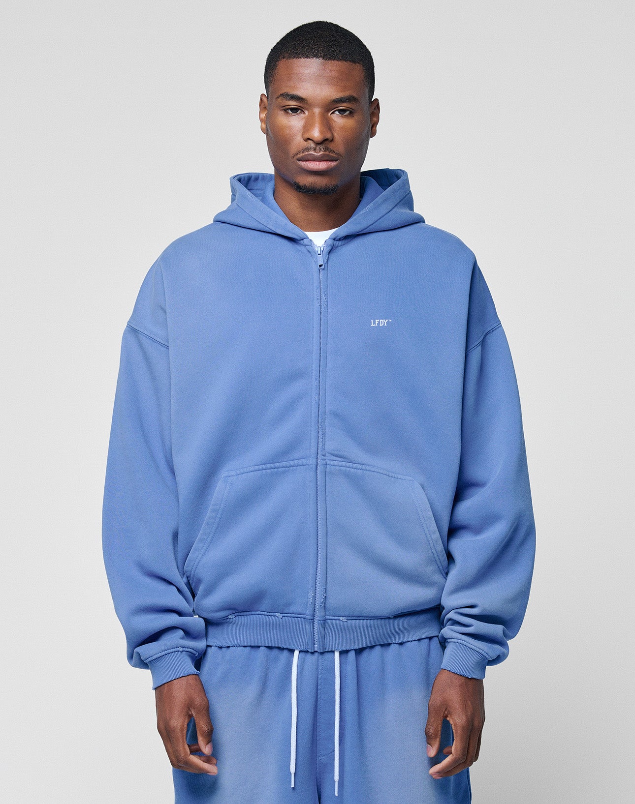 A man stands facing forward in a light blue LIVE FAST DIE YOUNG Basic Hooded Zip and matching sweatpants with an oversize fit, hands relaxed by his sides, against a plain light gray background.