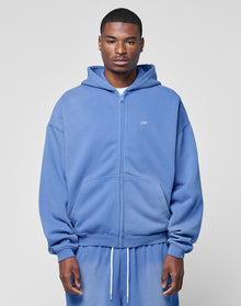 A man stands facing forward in a light blue LIVE FAST DIE YOUNG Basic Hooded Zip and matching sweatpants with an oversize fit, hands relaxed by his sides, against a plain light gray background.