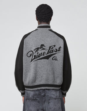 A person with short curly hair wears the LIVE FAST DIE YOUNG Boxy Knit Zip Jacket in gray and black with a galloping horse intarsia and “Live Fast Co.” on the back, paired with faded gray jeans, standing facing away from the camera.