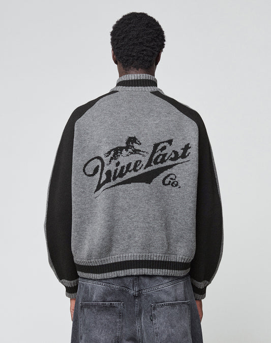 A person with short curly hair wears the LIVE FAST DIE YOUNG Boxy Knit Zip Jacket in gray and black with a galloping horse intarsia and “Live Fast Co.” on the back, paired with faded gray jeans, standing facing away from the camera.