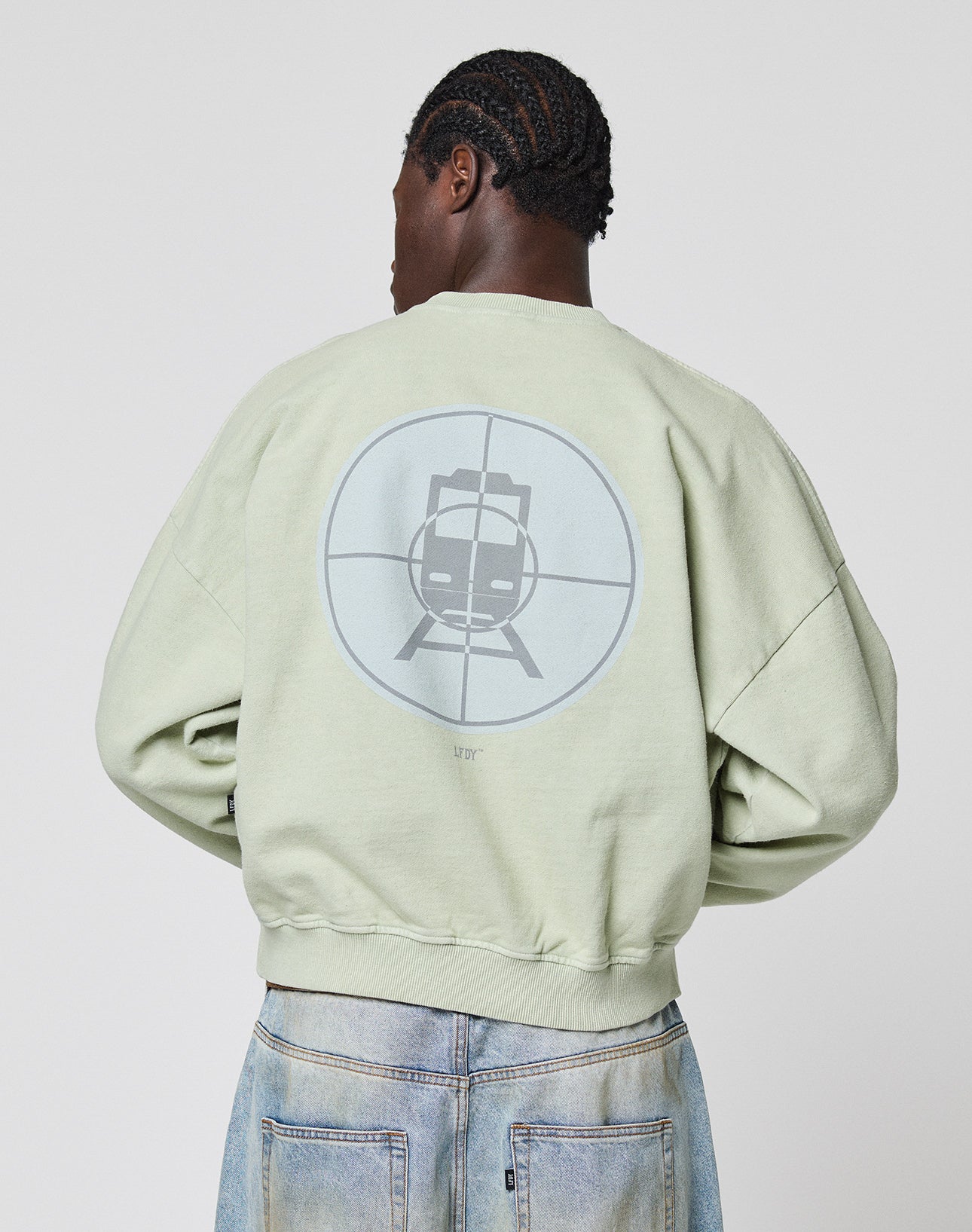 A person with braided hair, seen from behind, wears the light green LFDY Vandals Sweater by LIVE FAST DIE YOUNG, featuring an individueller Wasch-Effekt and a stylized train in a crosshair graphic on the back, paired with light blue jeans for an Oversize Fit.