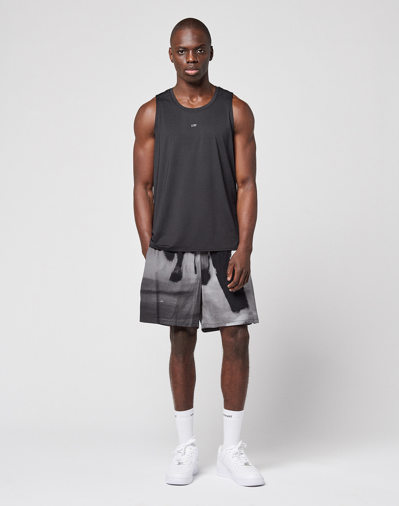 A man faces forward wearing the LFDY Performance Tank by LIVE FAST DIE YOUNG, featuring a reflective logo print, paired with black-and-white patterned Regular Fit shorts, white socks, and white sneakers against a light background.