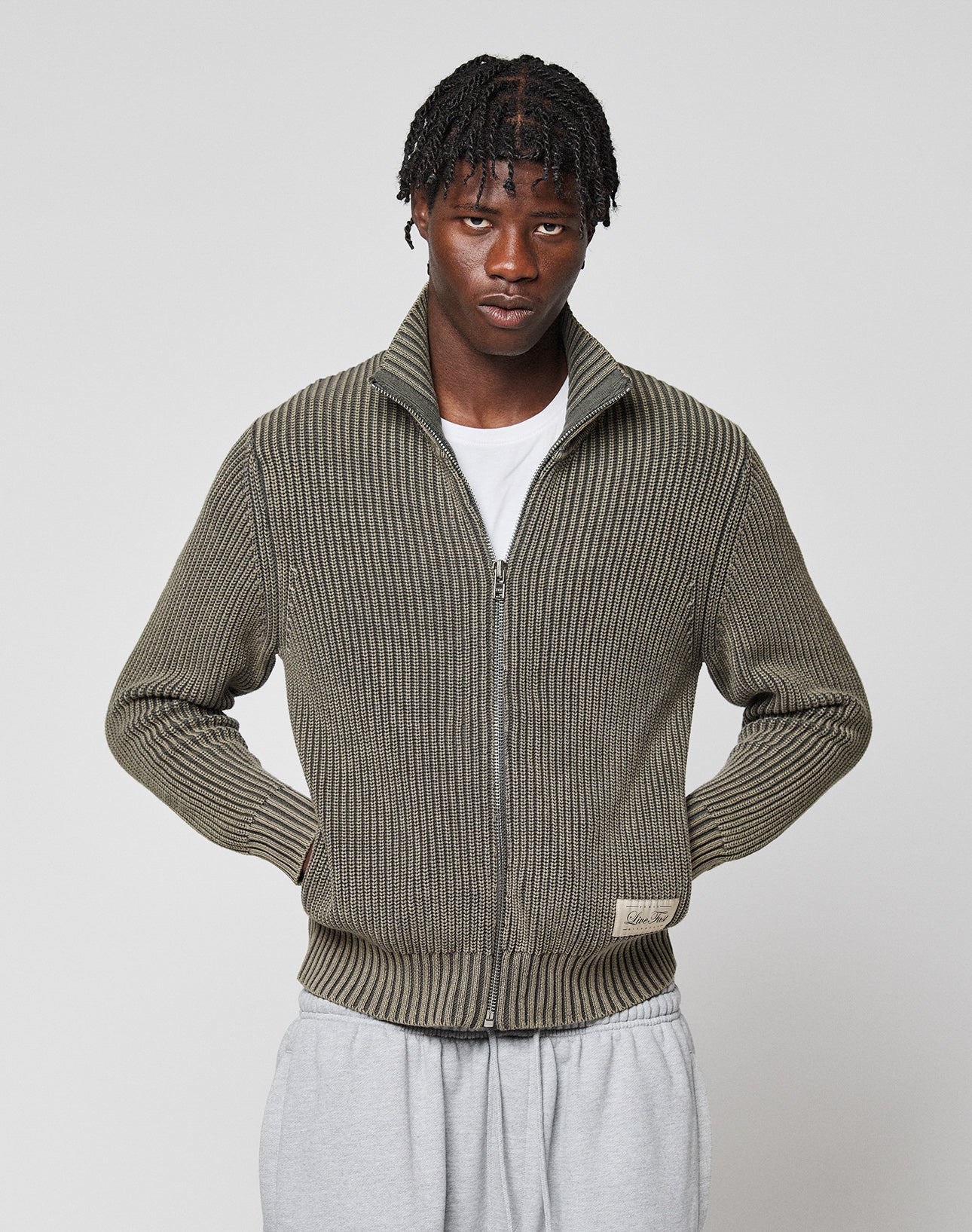 A person with short, braided hair wears an olive-green LFDY Knit Jacket by LIVE FAST DIE YOUNG over a white t-shirt and light gray sweatpants, standing against a plain light gray background.