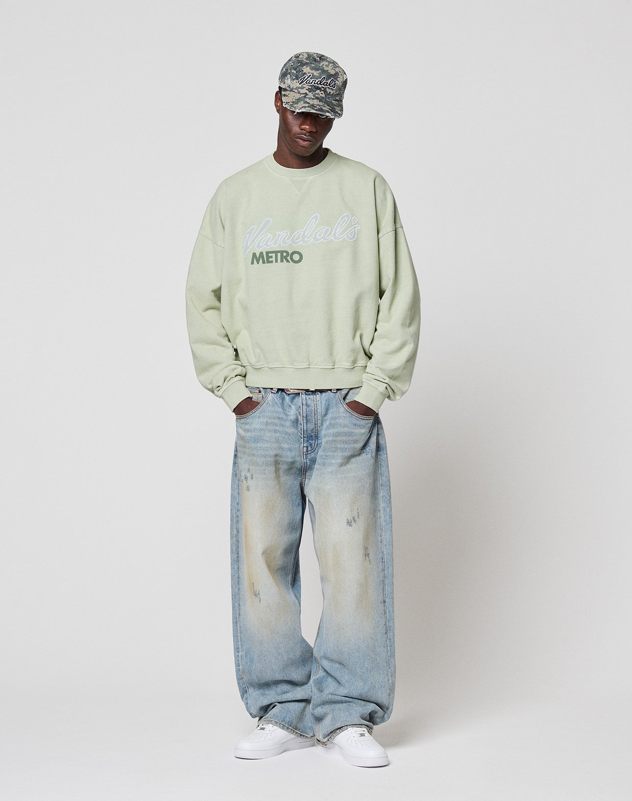 A person in an Oversize Fit camouflage cap, light green LIVE FAST DIE YOUNG LFDY Vandals Sweater with unique wash effect, baggy light blue jeans, and white sneakers stands against a plain white background.