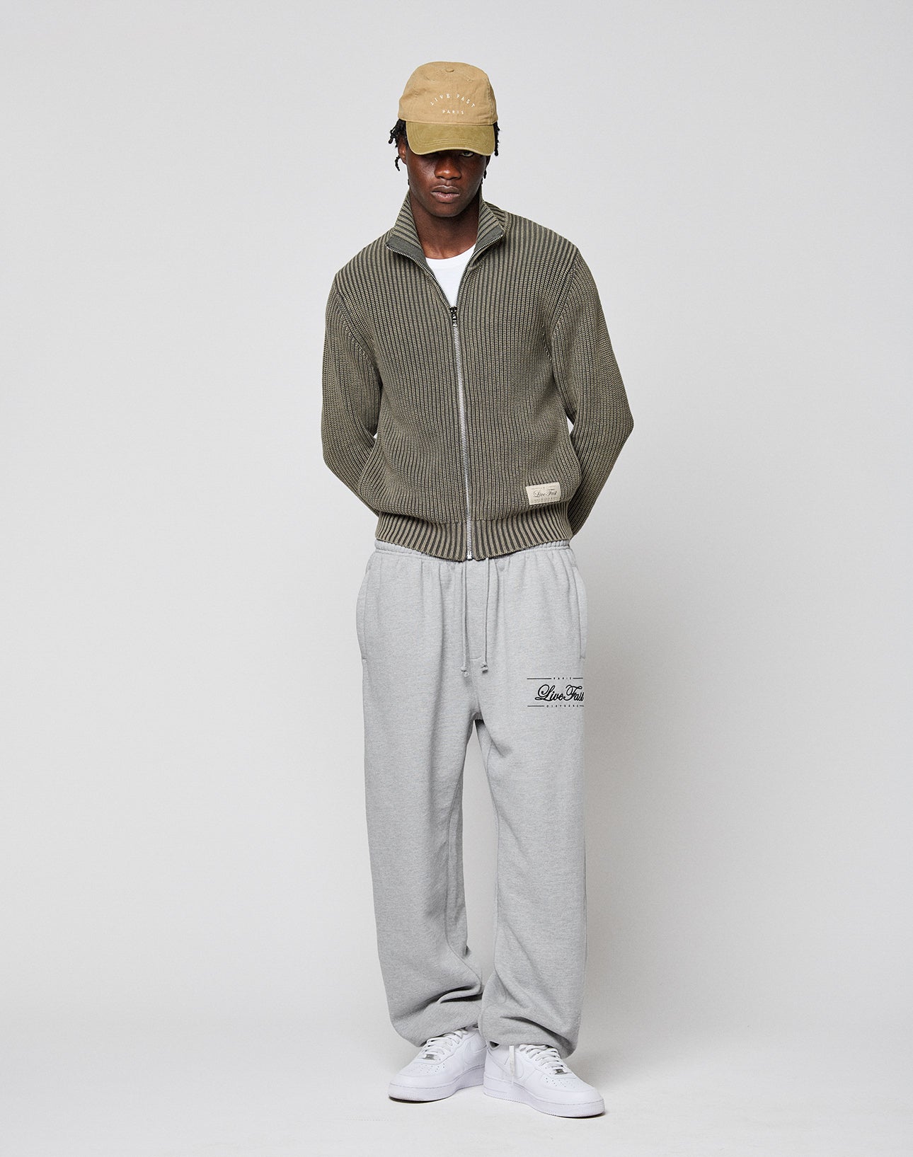 A person in a beige cap, LFDY Knit Jacket by LIVE FAST DIE YOUNG, light gray sweatpants, and white sneakers stands against a plain light background with hands in pockets, looking slightly downward.