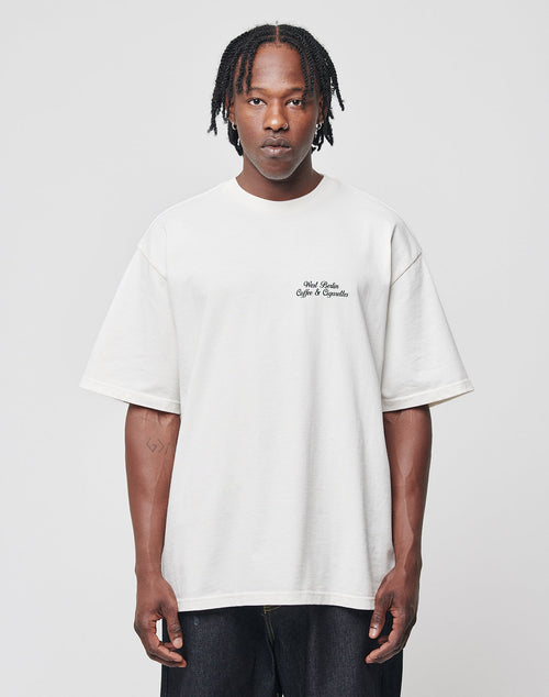 A man with braided hair wears the LIVE FAST DIE YOUNG Live Fast Coffee Tee, an oversize white 100% Baumwolle T-shirt featuring small black text on the left chest, standing against a plain light gray background with a neutral expression.