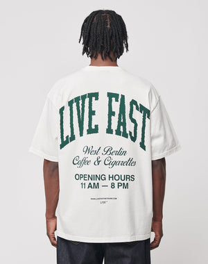 A person faces away from the camera, wearing the LIVE FAST DIE YOUNG Live Fast Coffee Tee—an oversize fit white T-shirt made of 100% cotton with green and black back text: “LIVE FAST, LFDY Berlin Café & Cigarettes, OPENING HOURS 11 AM – 8 PM.”.