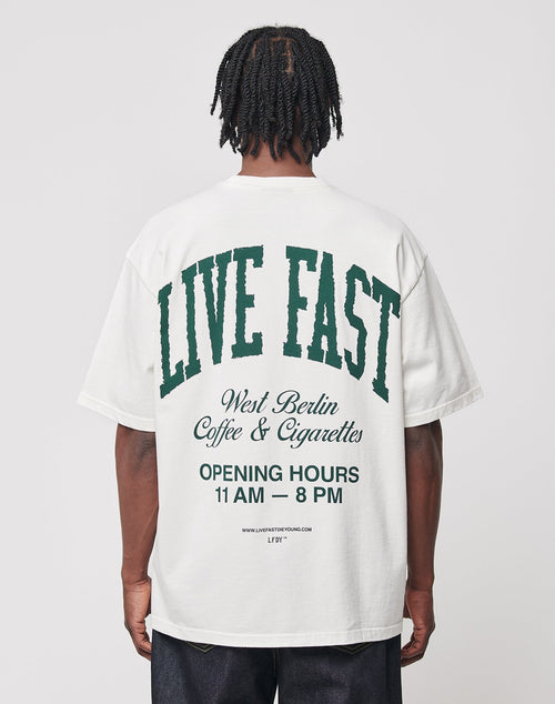 A person faces away from the camera, wearing the LIVE FAST DIE YOUNG Live Fast Coffee Tee—an oversize fit white T-shirt made of 100% cotton with green and black back text: “LIVE FAST, LFDY Berlin Café & Cigarettes, OPENING HOURS 11 AM – 8 PM.”.