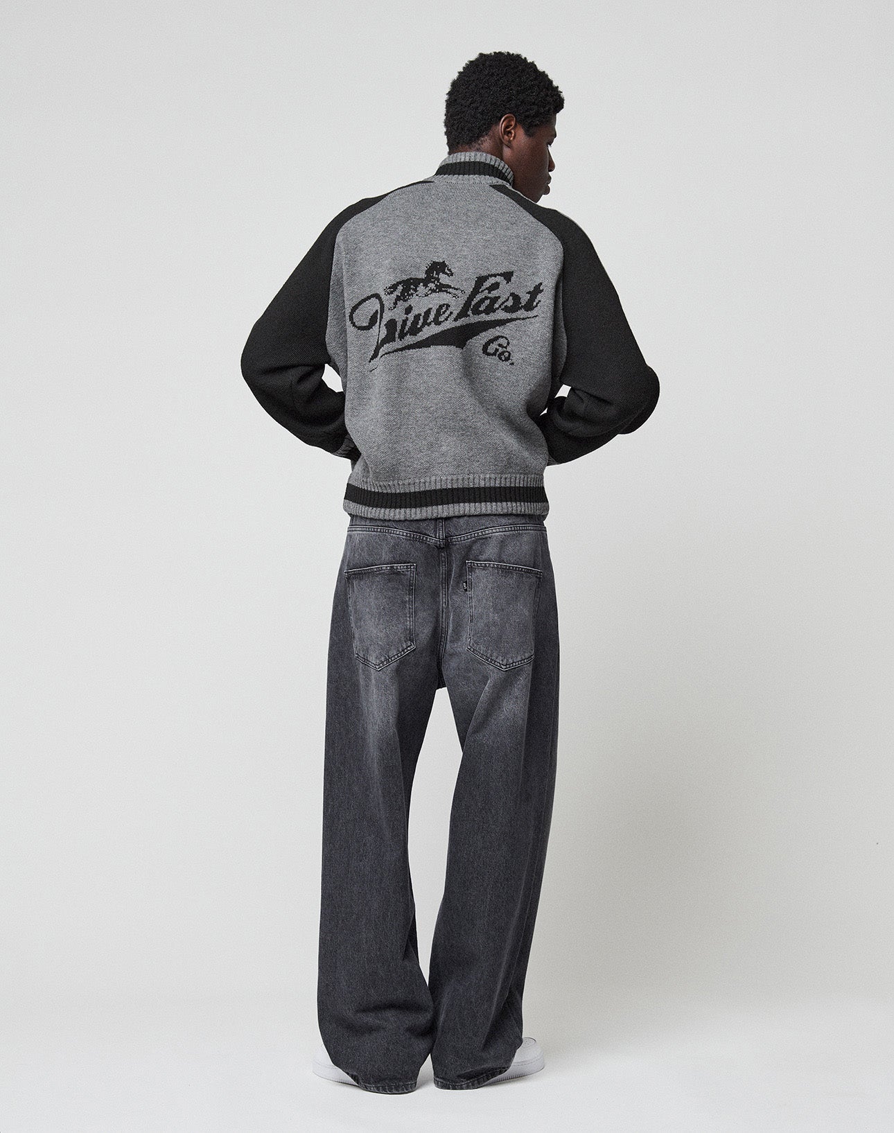 A person stands facing away in a LIVE FAST DIE YOUNG Boxy Knit Zip Jacket with gray and black colors, an intarsia-knit horse graphic on the back, loose dark gray jeans, and white shoes against a plain light background.