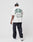 A person in a white hat, dark baggy jeans, and white sneakers faces away against a plain white background, wearing the LIVE FAST DIE YOUNG Live Fast Coffee Tee—an oversize fit white T-shirt with bold green text and Berlin Café details.