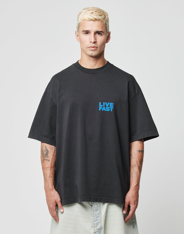 A person with short bleached blond hair wears the LFDY 4 Ever Tee from LIVE FAST DIE YOUNG—an oversized black cotton t-shirt with LIVE FAST in blue on the chest, standing against a plain light background with tattoos visible on both arms.