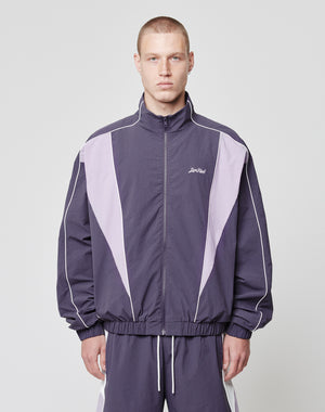 A person stands facing forward against a light gray background, wearing the LIVE FAST DIE YOUNG Basic Nylon Trackjacket in purple and lavender with matching pants. The regular fit features white piping, side seam pockets, and a Lost Art logo on the left chest.