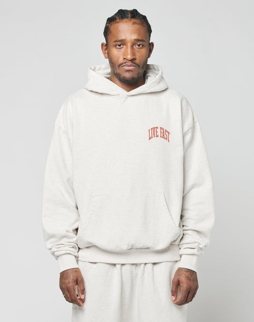 A man faces forward in a light beige 100% Baumwolle Live Fast Amsterdam Hooded by LIVE FAST DIE YOUNG, paired with matching sweatpants. An oversize fit, it sports LIVE FAST in red on the chest. The background is plain and light-colored.