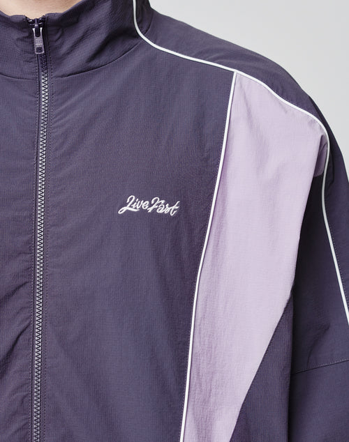 A close-up of a person wearing the LIVE FAST DIE YOUNG Basic Nylon Trackjacket in purple with light lavender and white accents. The regular fit has side entry pockets and Live Fast embroidered in white script on the left chest.